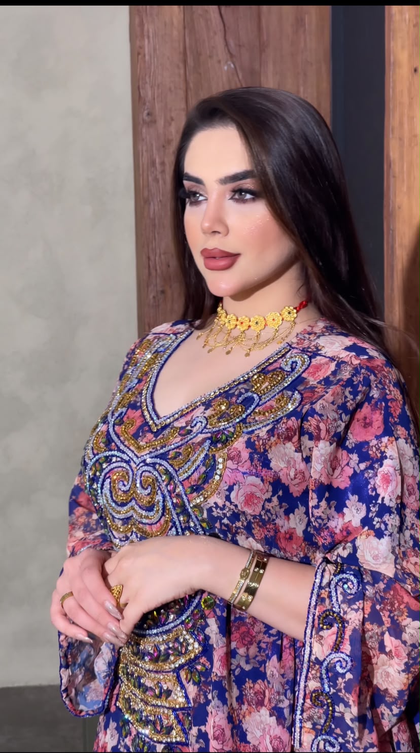 قفطان طويل بأكمام واسعة، مصنوع من قماش أزرق ملكي فاخر بنقوش زهرية وردية وبيضاء كثيفة.